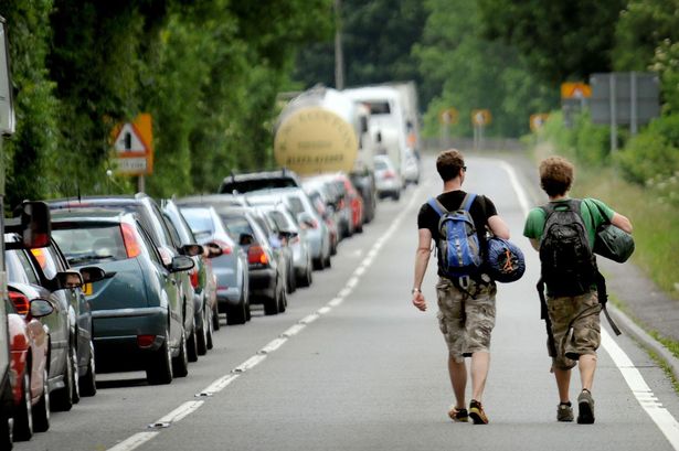 queue of cars along the left hand side of a road with two festival goers walking alongside the right